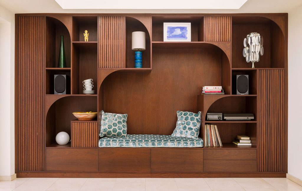 Dark wood bookcase with open shelves, cabinets and drawers including some slatted cupboard doors. The shelves are geometric with a curved detail motif. The cabinet displays art, houses a hifi with record collection, it also has a bar and an upholstered seating area.
